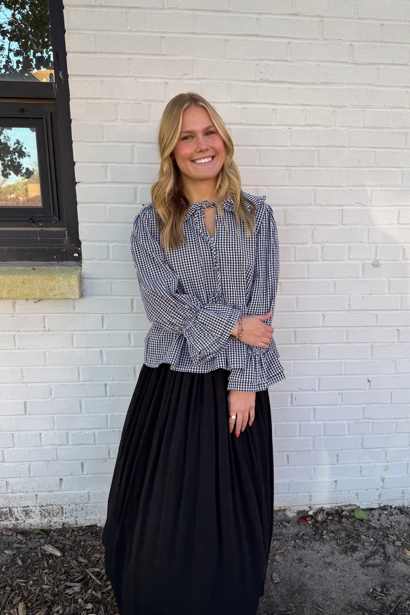 Woman wearing a checkered blouse and black skirt standing against a white brick wall.