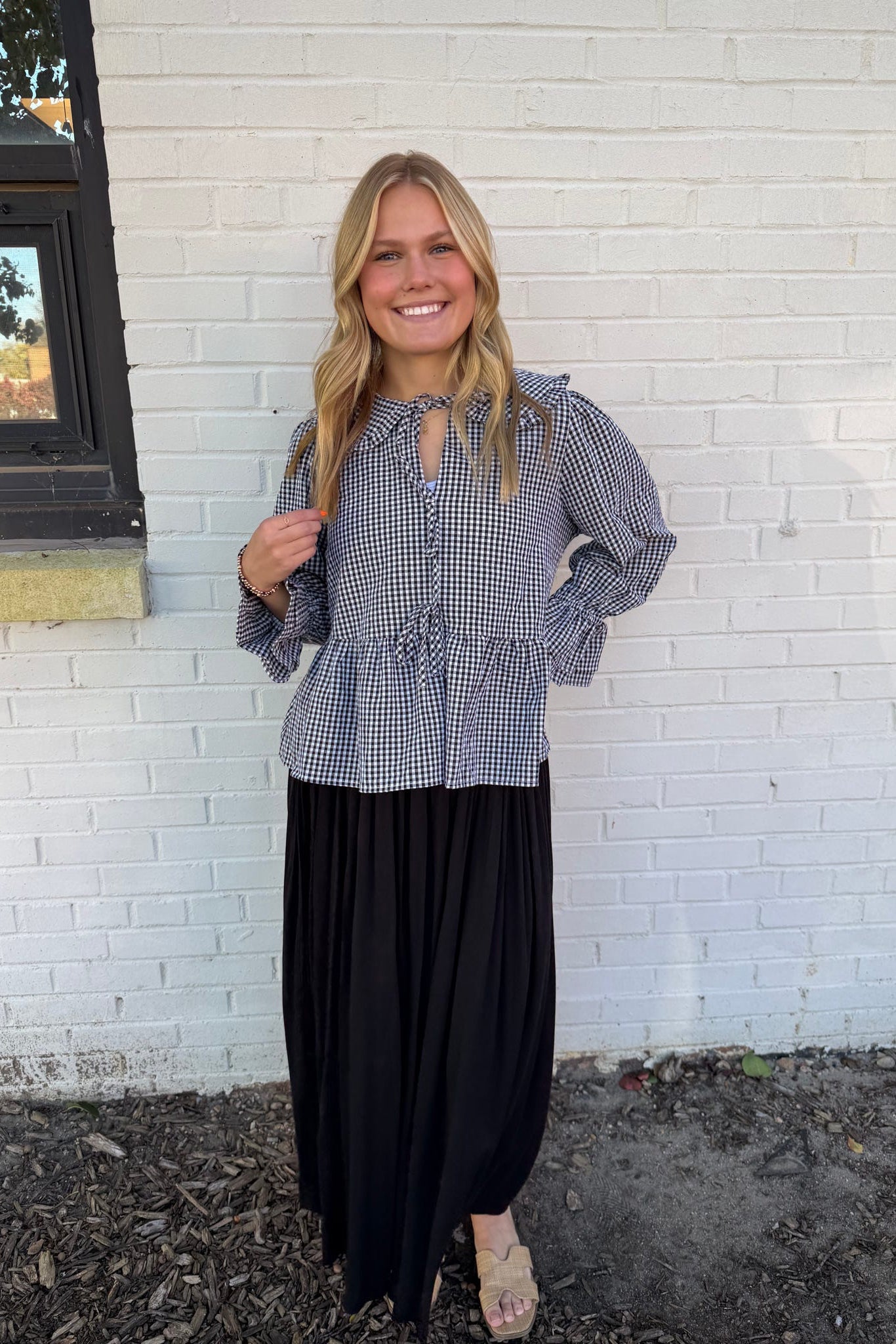 Woman wearing a checkered blouse and black skirt standing in front of a white brick wall.