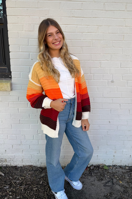 Woman wearing a colorful striped sweater and blue jeans standing against a white brick wall.