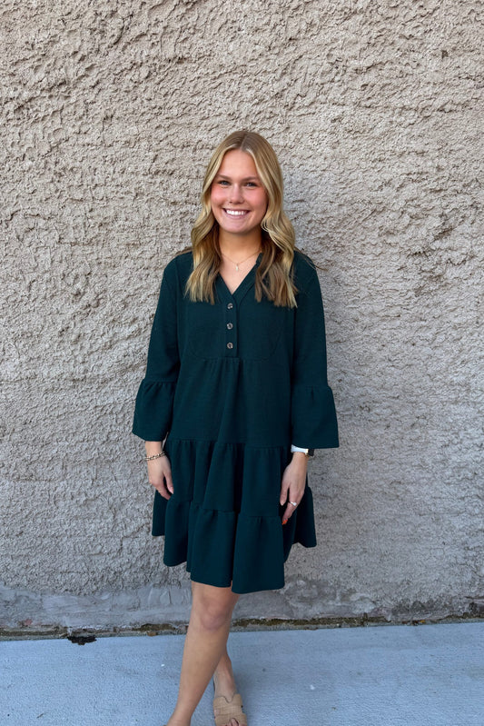 Woman wearing a dark green dress standing against a textured wall.