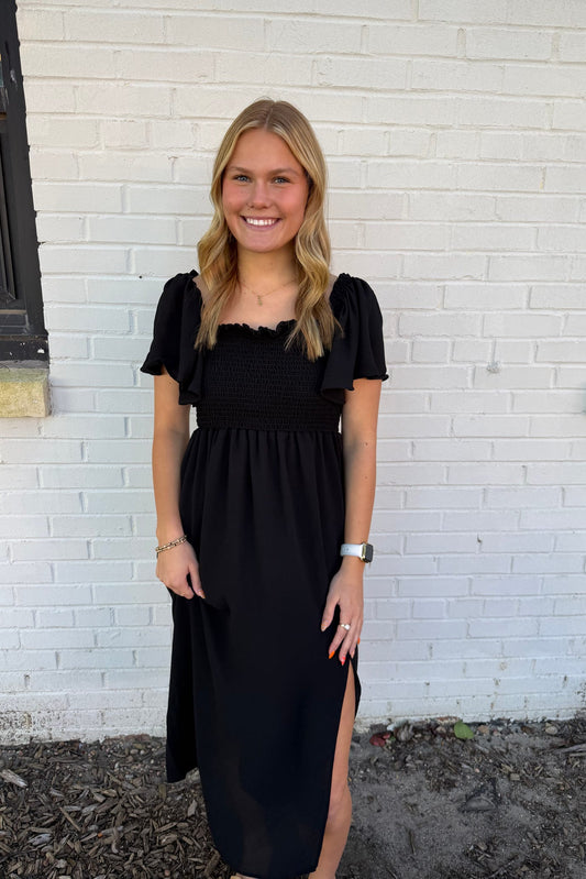 Woman wearing a black dress standing against a white brick wall.