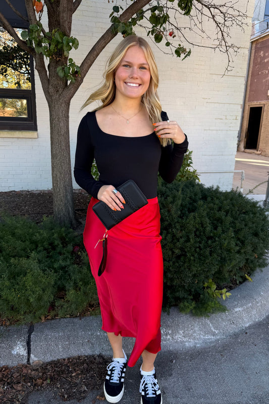 Woman in black top and red skirt standing outdoors with a building and tree in the background