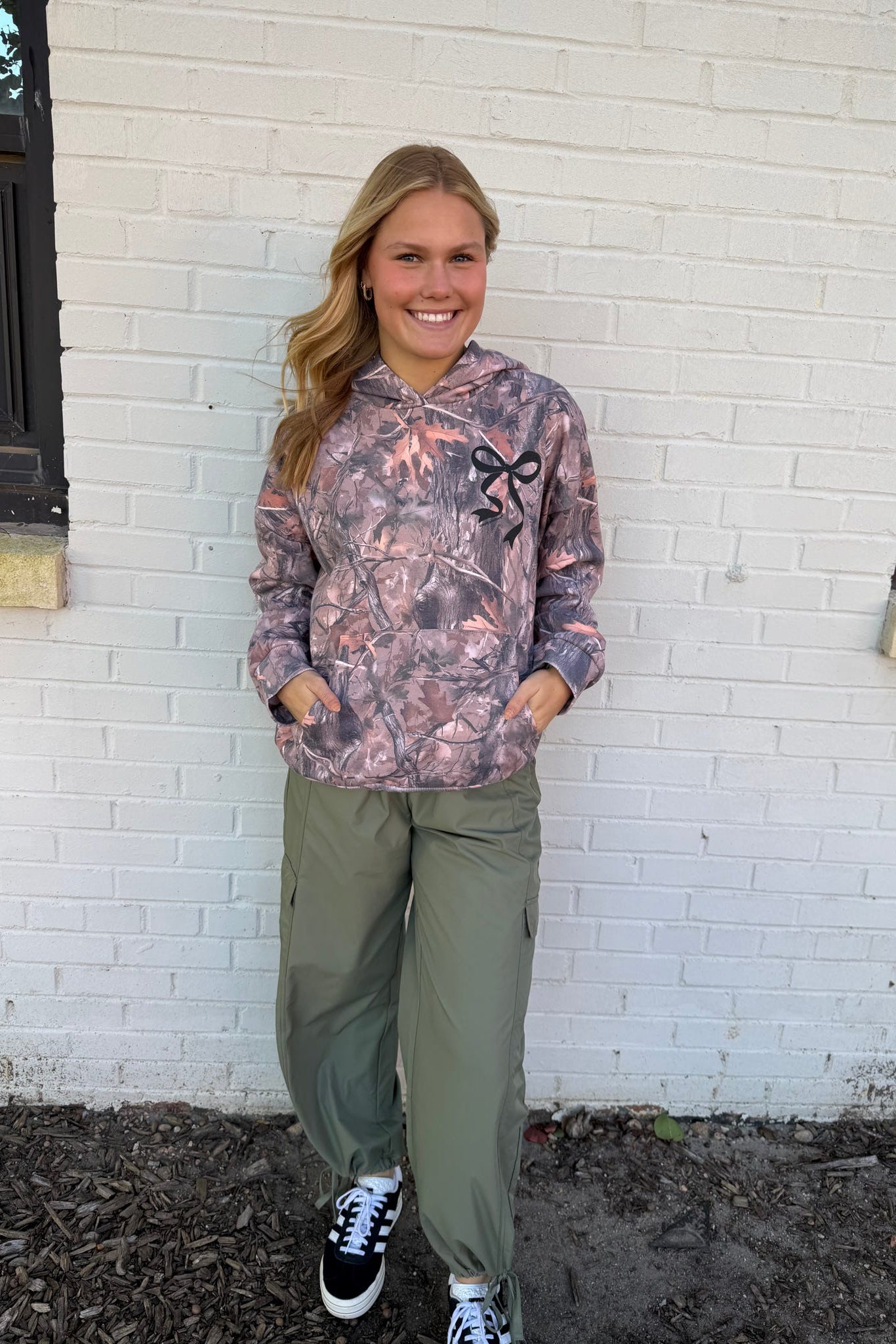 Woman wearing a patterned hoodie and green pants standing in front of a white brick wall.