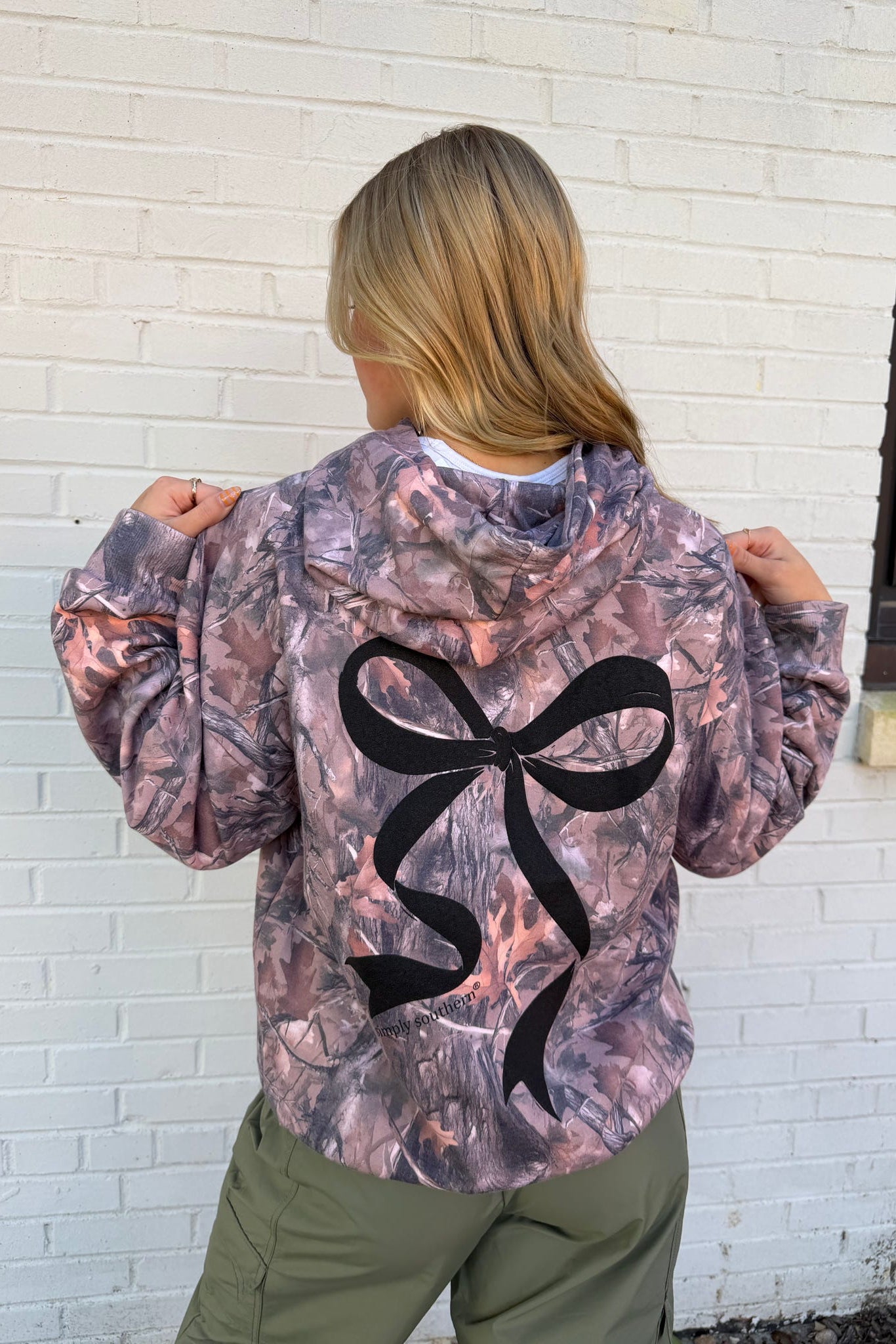 Person wearing a camouflage jacket with a black bow design on the back, standing against a white brick wall.