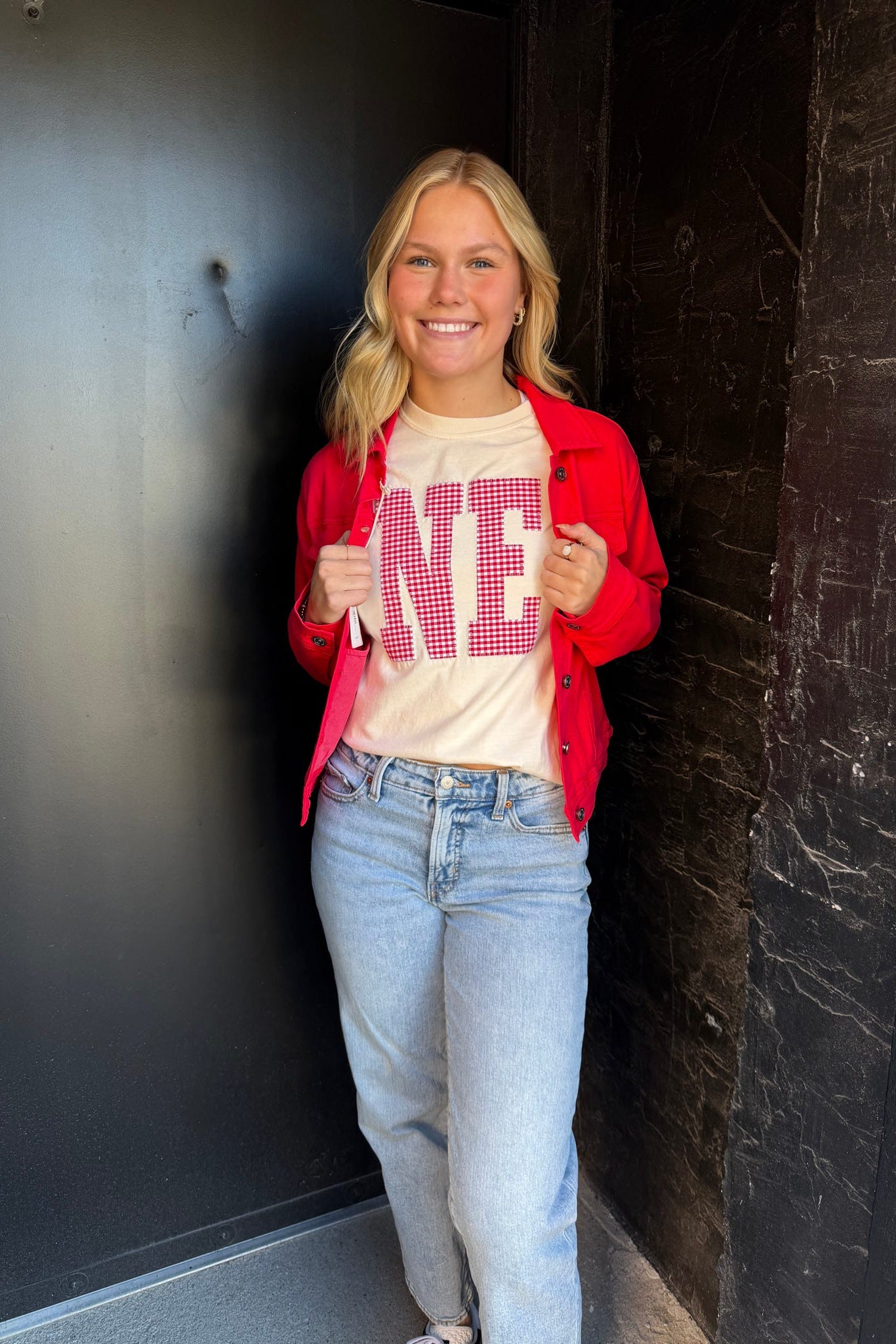 Person wearing a red jacket, white t-shirt with text, and blue jeans standing against a dark wall.