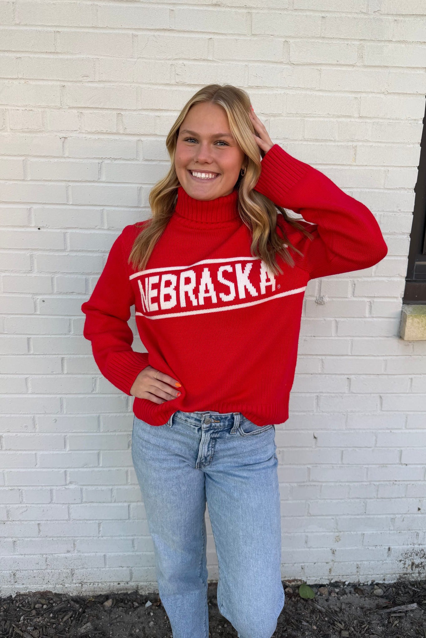 Person wearing a red 'NEBRASKA' sweater against a white brick wall.