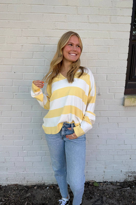 Woman wearing a yellow and white striped sweater and blue jeans standing against a white brick wall.
