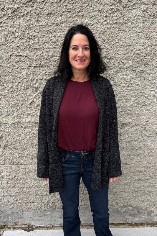 Woman wearing a dark cardigan, burgundy shirt, and jeans standing against a textured wall.