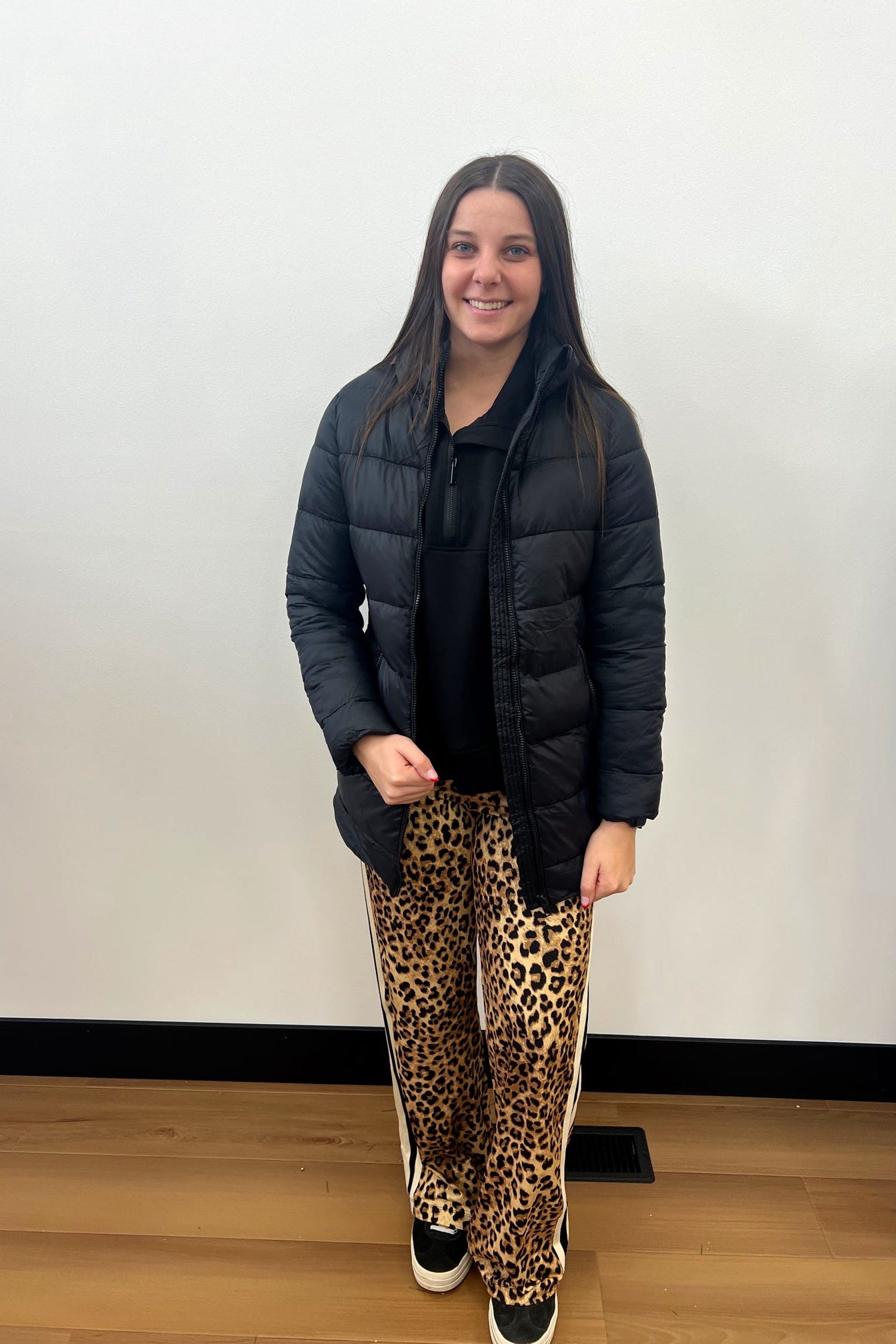 Person wearing a black puffer jacket, black top, and leopard print pants standing against a white wall.