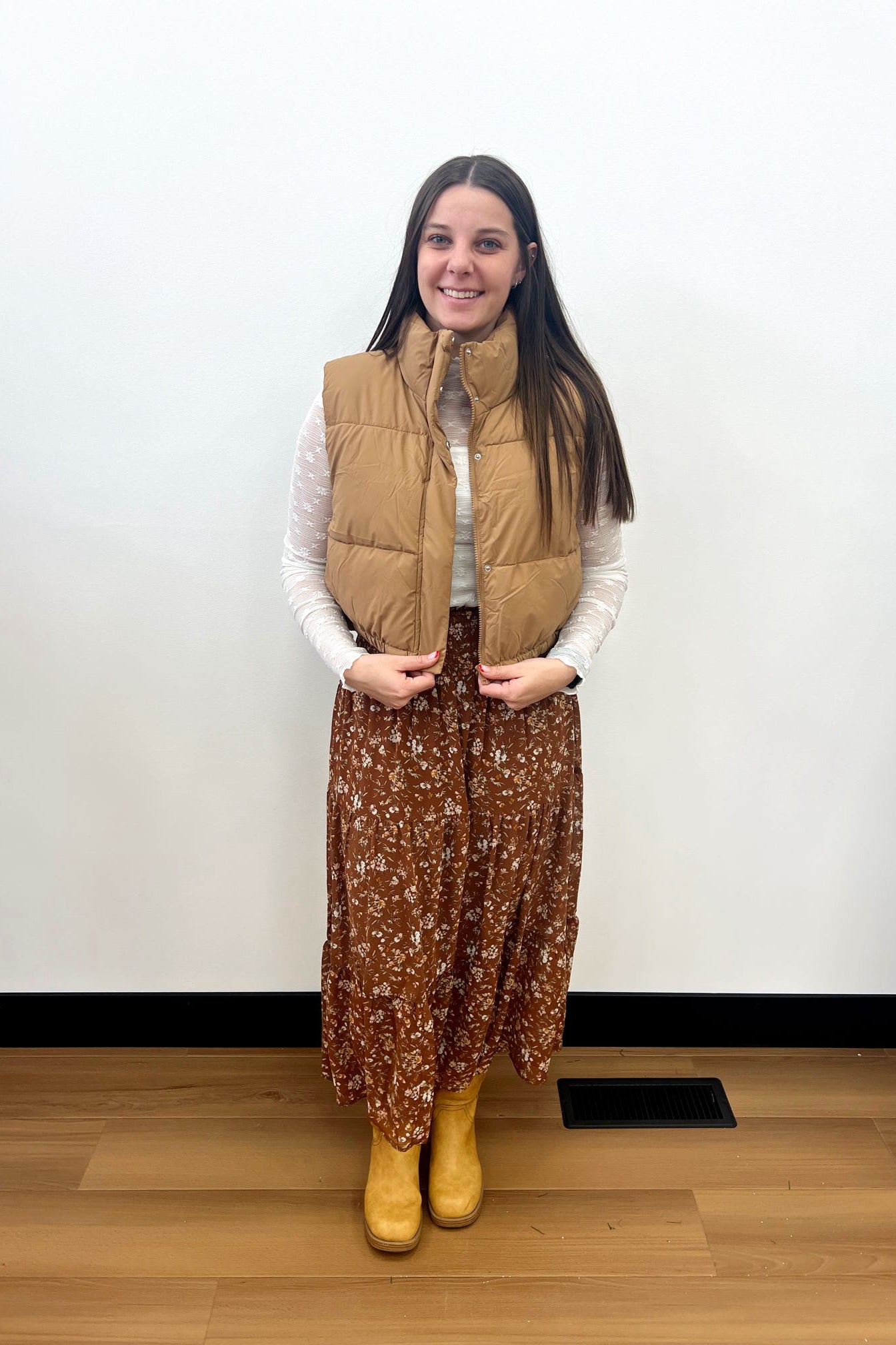 Person wearing a tan vest, white shirt, brown floral skirt, and yellow boots against a white wall.