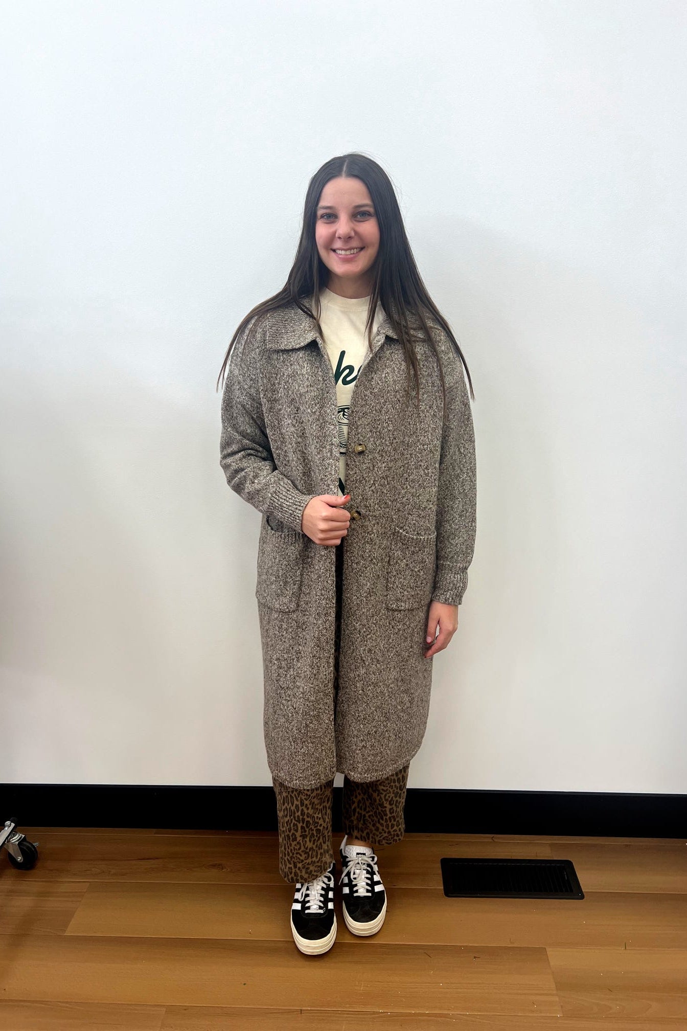 Person wearing a long brown coat standing against a white wall.