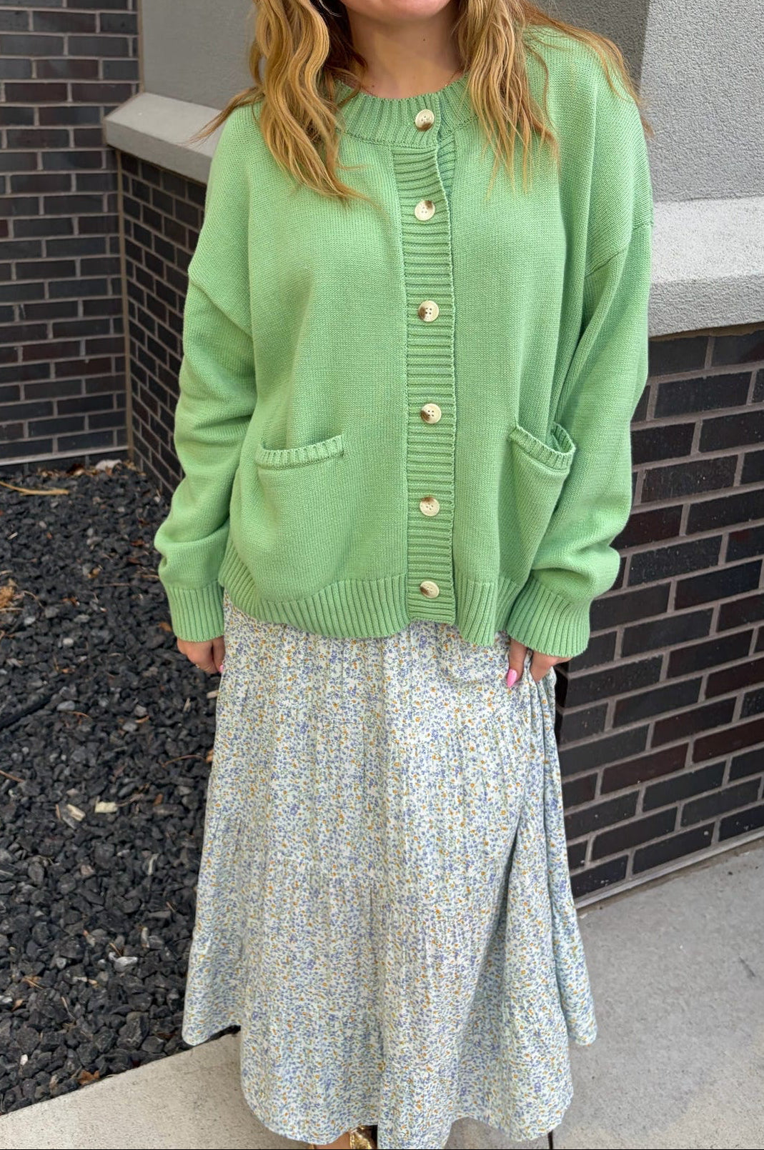 Woman wearing a green cardigan and light-colored dress standing against a brick wall.