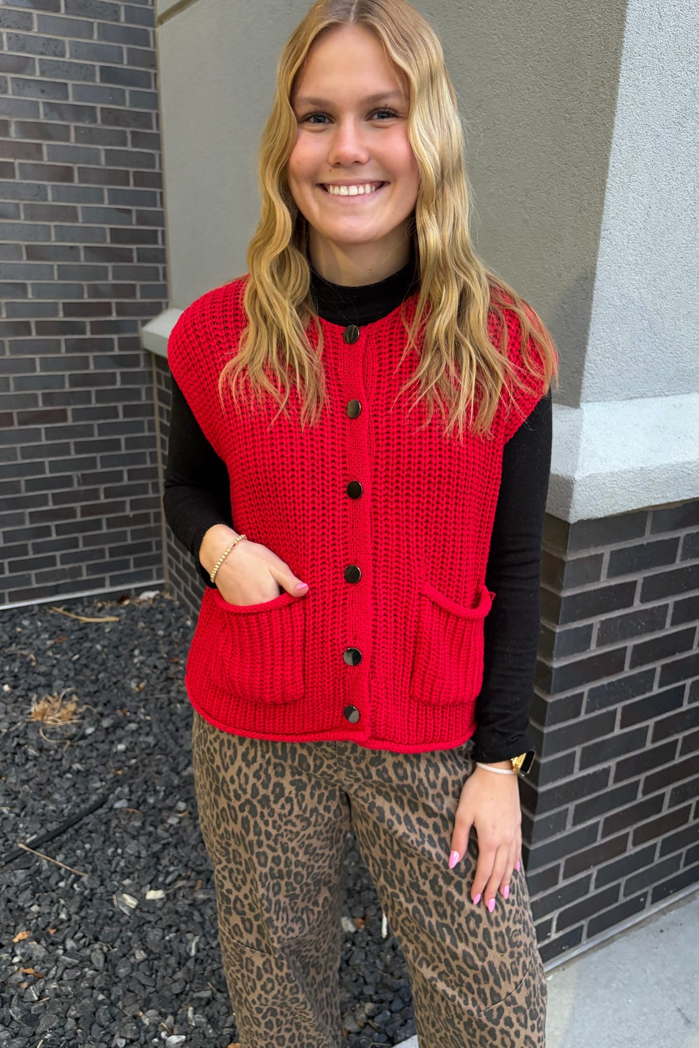 Person wearing a red vest over a black shirt with leopard print pants, standing against a brick wall.