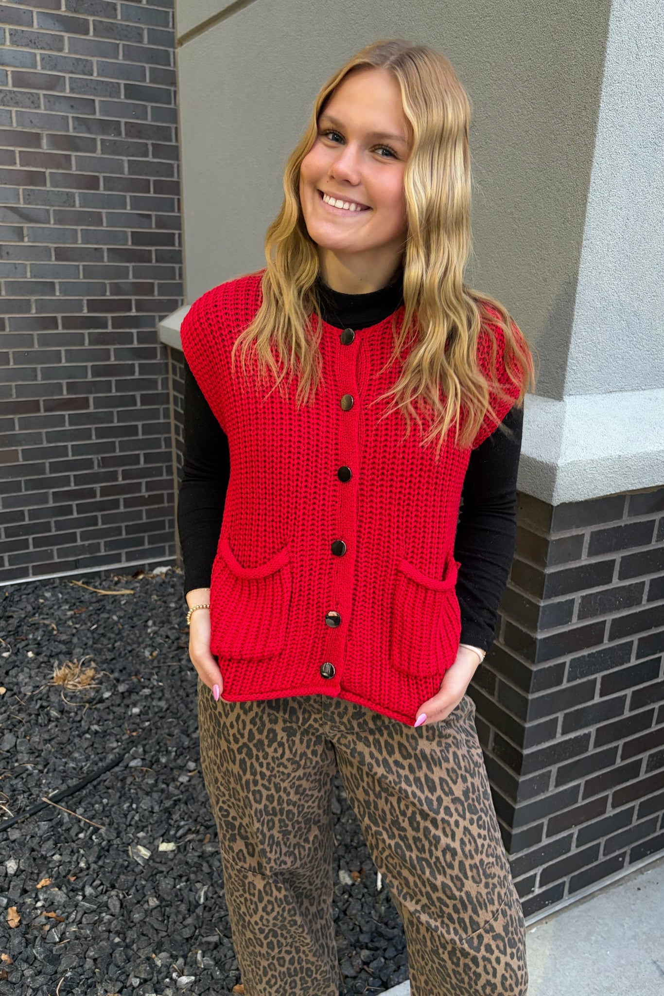 Person wearing a red knitted vest and leopard print pants standing against a brick wall.