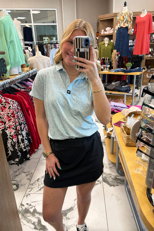 Woman taking a mirror selfie in a clothing store wearing a light blue polo shirt and black skirt.