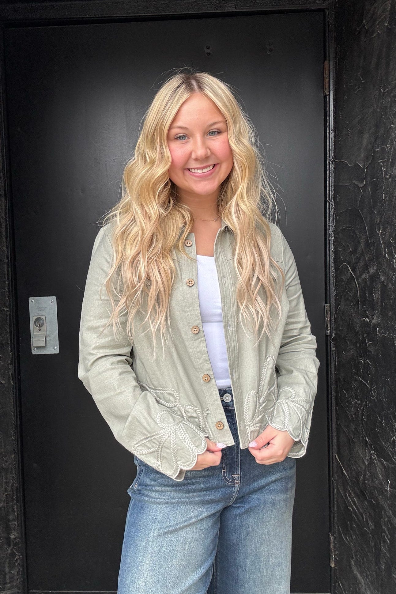 Woman wearing a light green jacket and blue jeans standing in front of a black door.