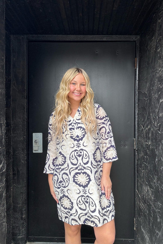 Woman wearing a white dress with blue patterns standing in front of a black door.