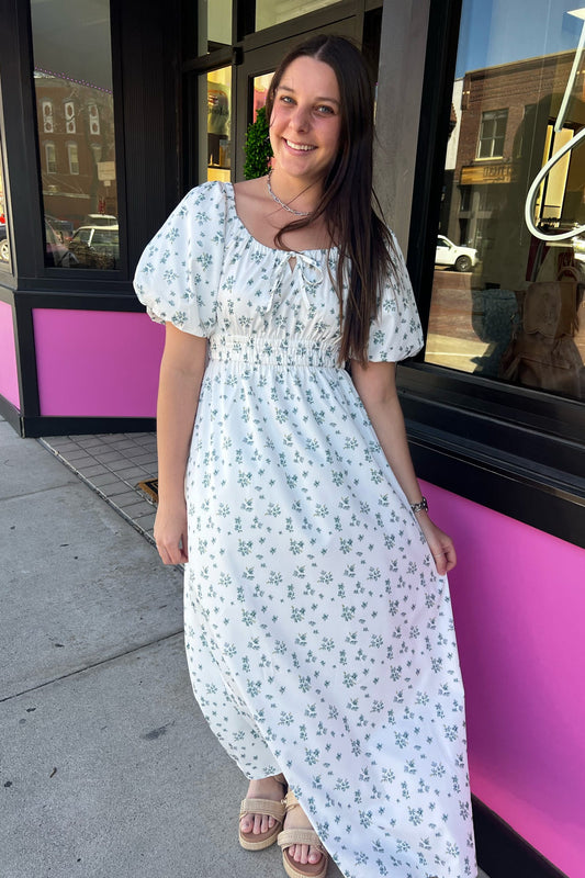 Woman in a white floral dress standing on a sidewalk with a pink building in the background