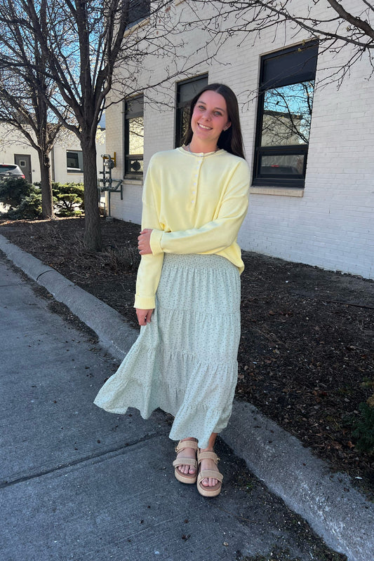 Woman in a yellow top and light blue skirt standing on a sidewalk with a building in the background.