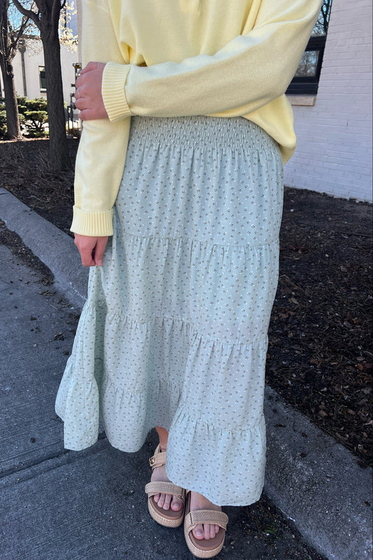 Person wearing a yellow sweater and light green skirt on a sidewalk.