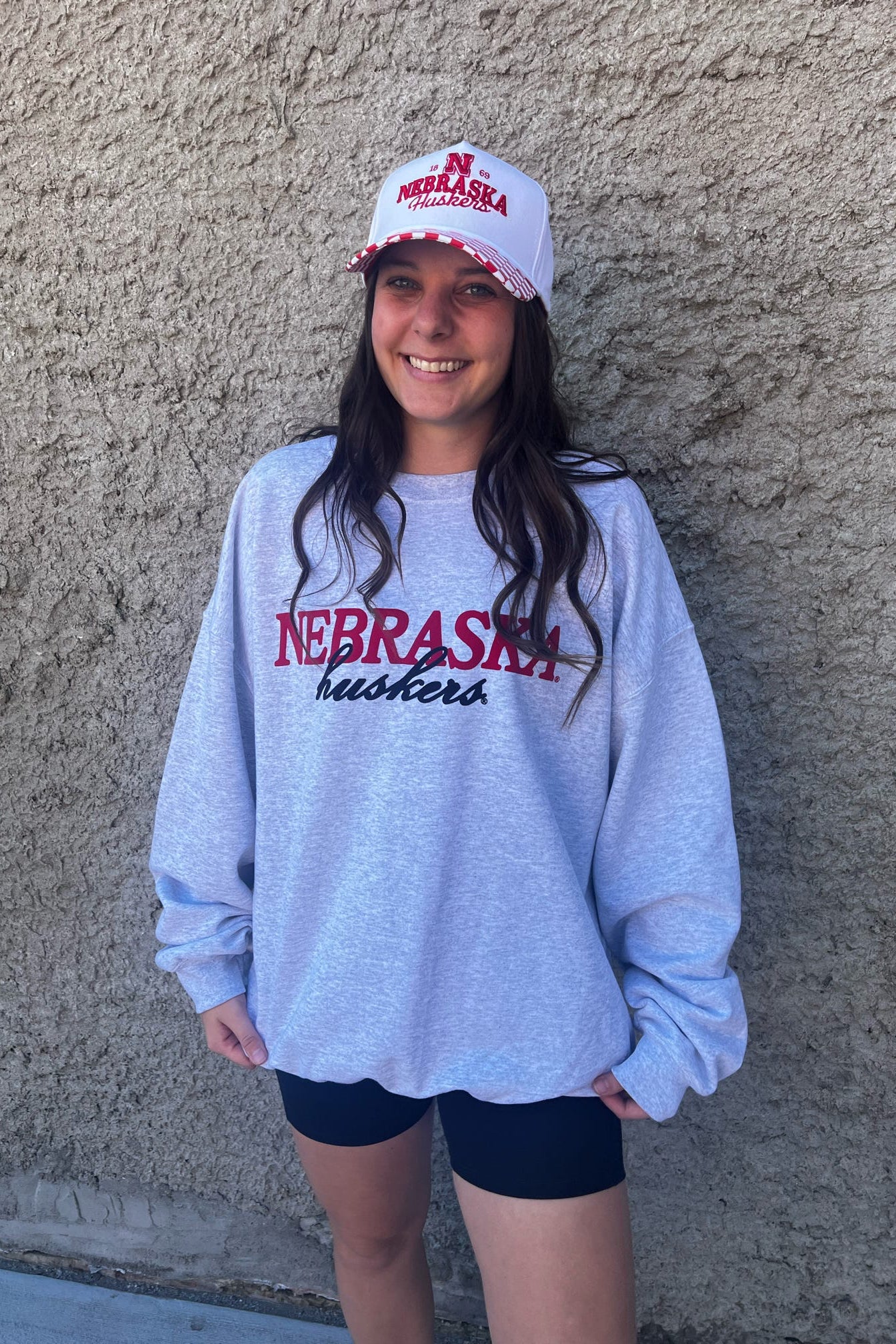 Person wearing a Nebraska shirt and cap against a concrete wall