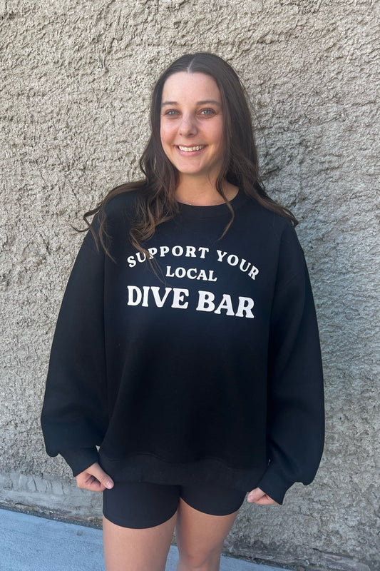 Person wearing a black sweatshirt with 'Support Your Local Dive Bar' text against a gray wall.