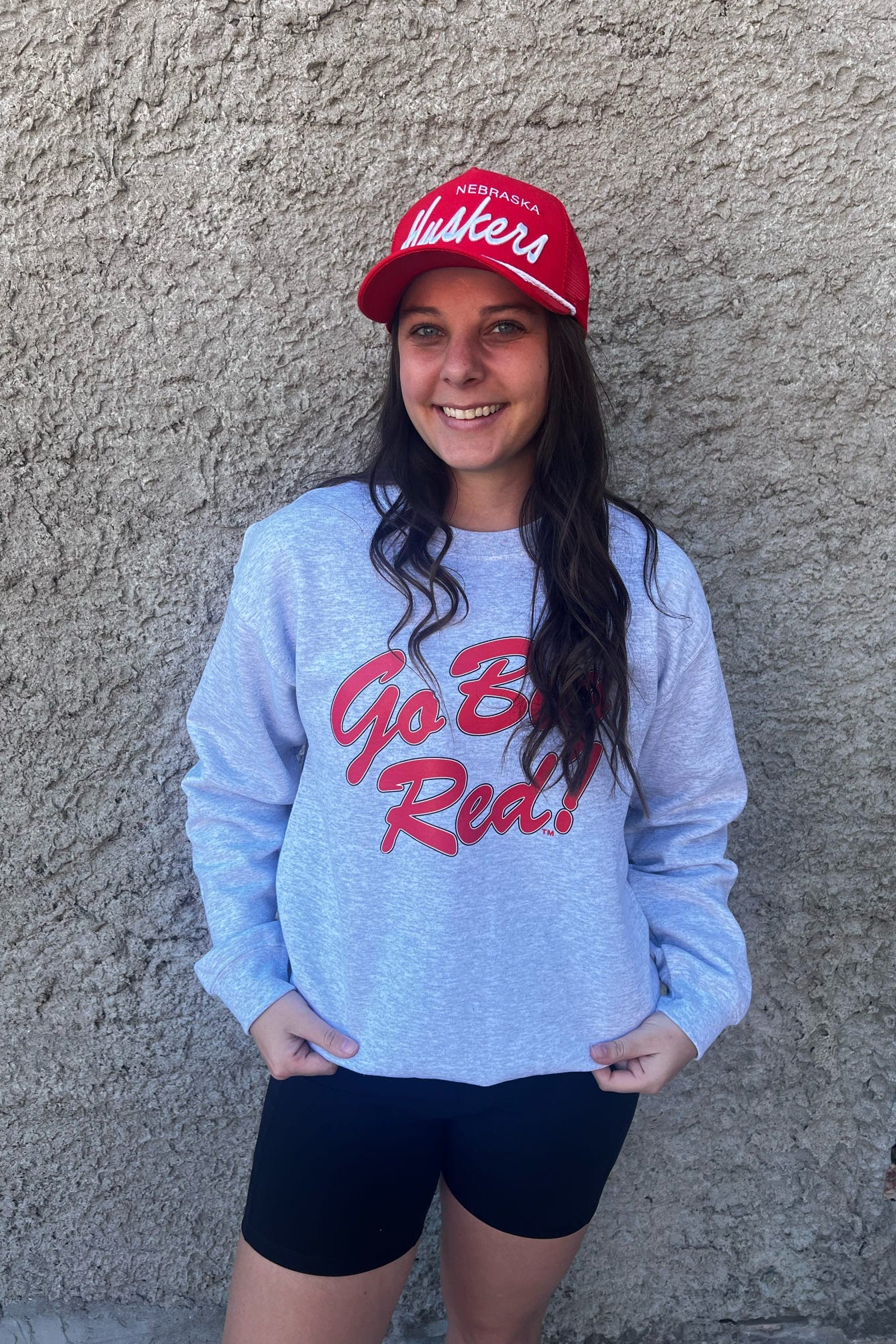 Person wearing a blue sweatshirt with red text and a red cap, standing against a textured wall.