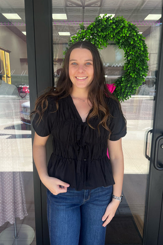 Woman standing in front of a glass door with a wreath, wearing a black top and blue jeans.