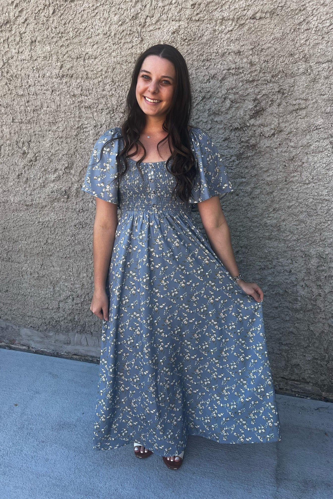 Woman in a blue floral dress standing against a textured wall.