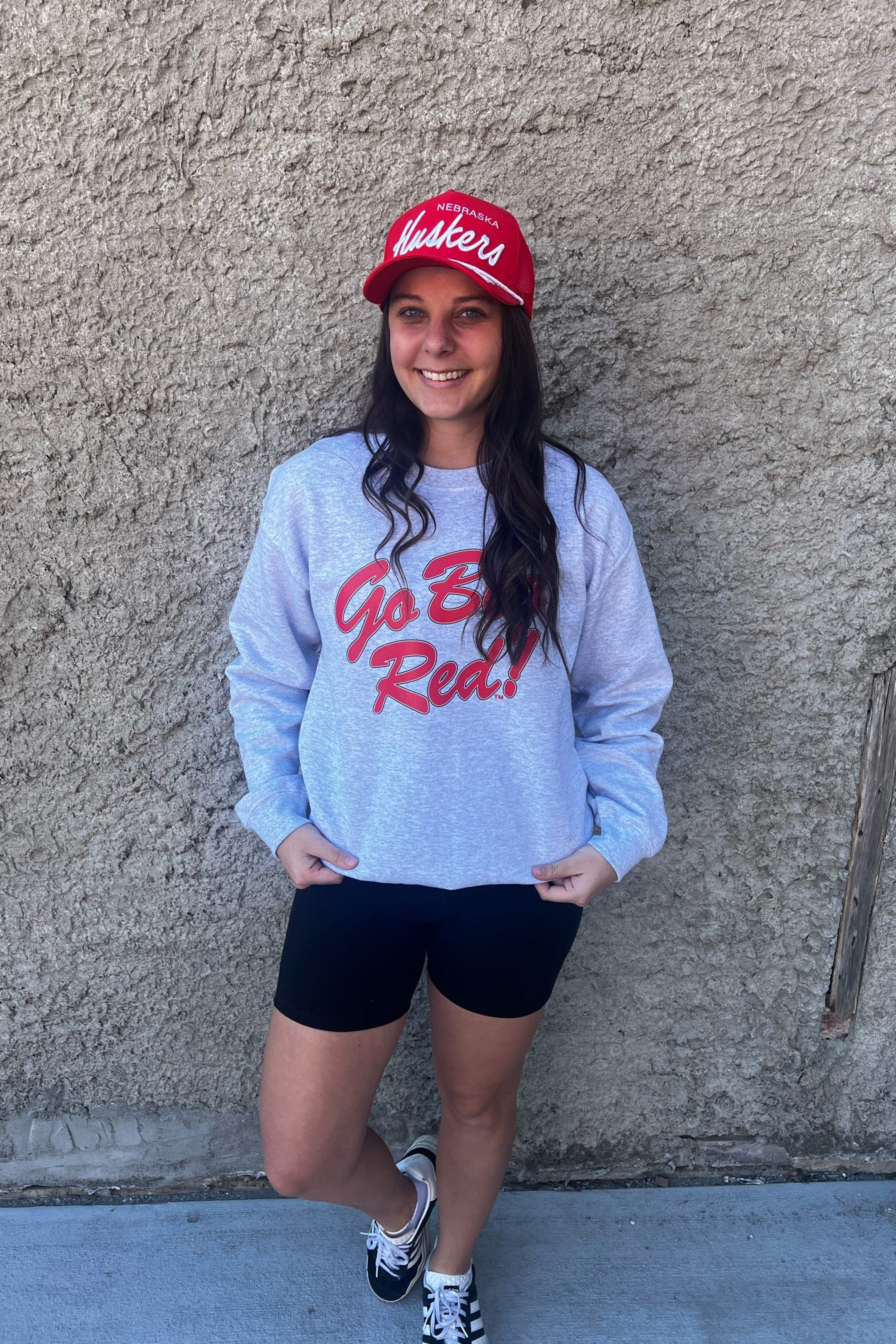 Person wearing a gray sweatshirt with red text and a red cap, standing against a beige wall.