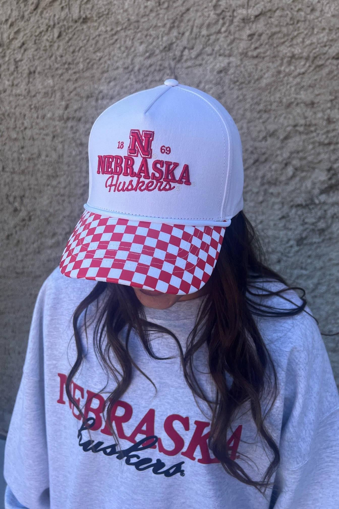 Person wearing a Nebraska Huskers cap and sweatshirt against a beige wall