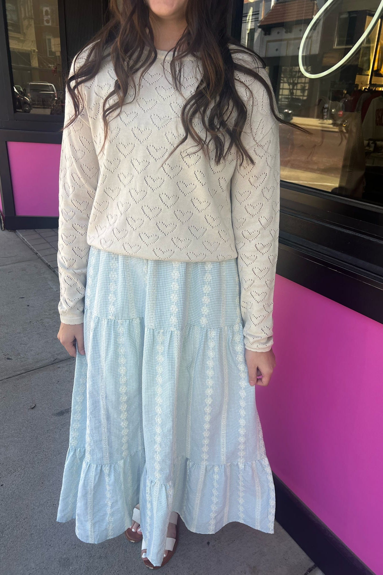 Woman wearing a light-colored lace top and light blue tiered skirt standing in front of a pink building.