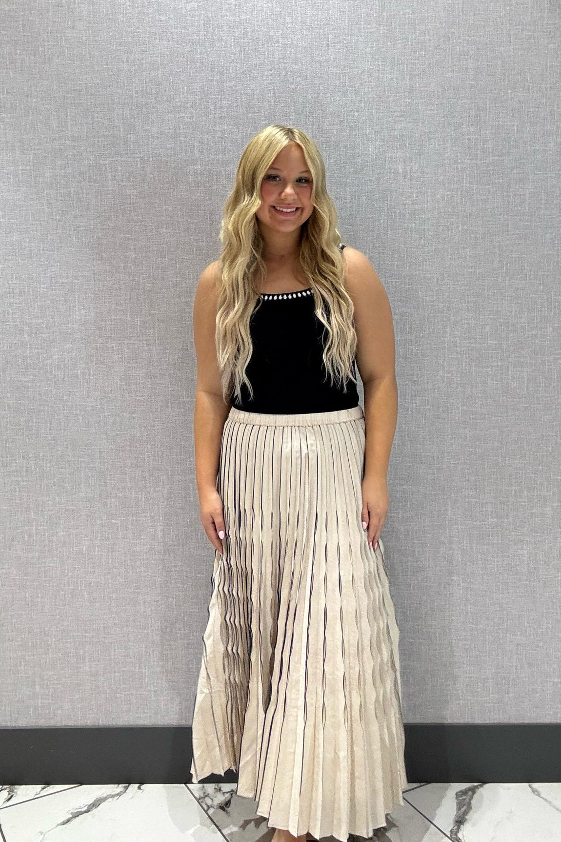 Woman wearing a navy top and beige pleated skirt standing against a gray wall.