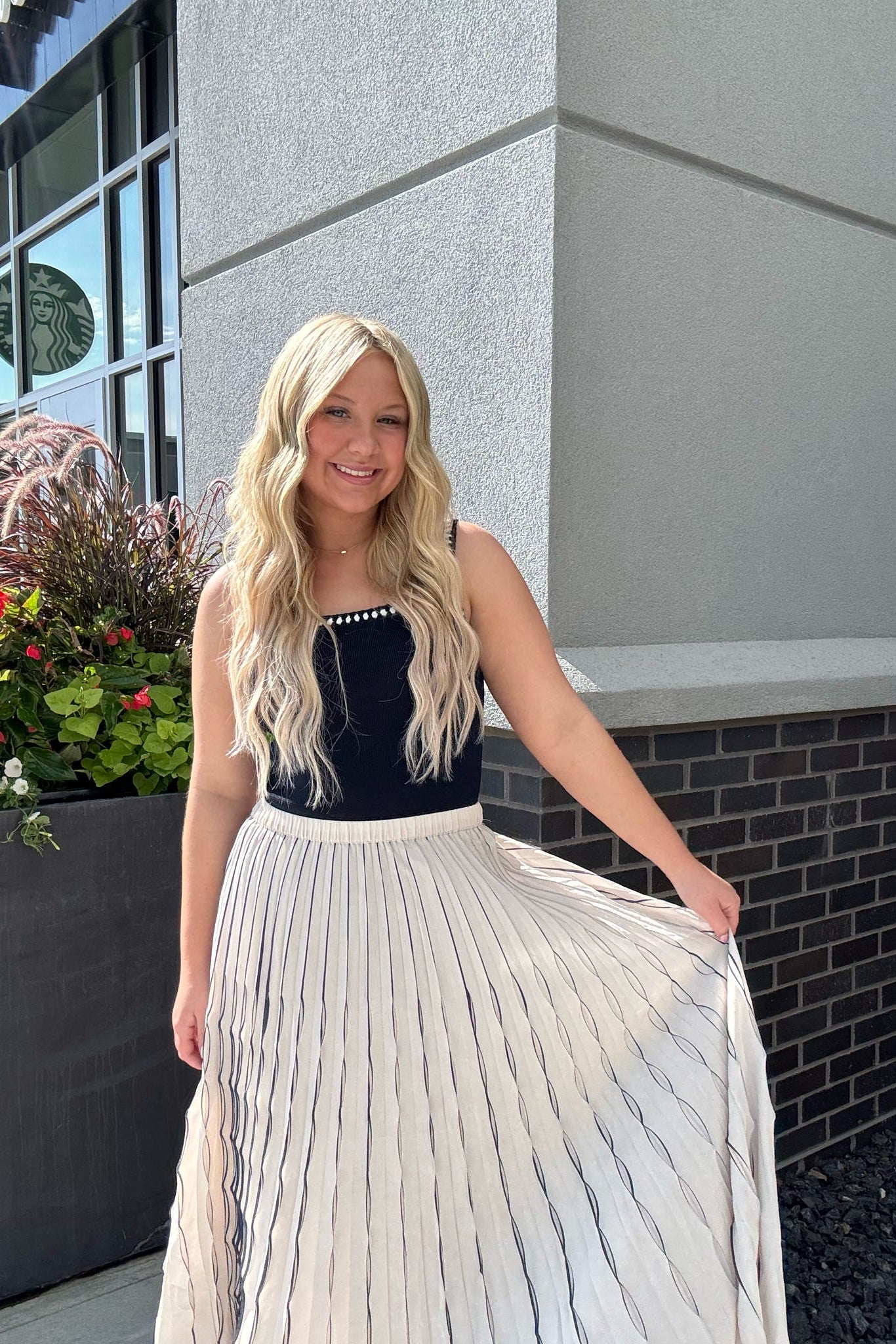 Woman in a navy top and white pleated skirt standing in front of a building with plants.