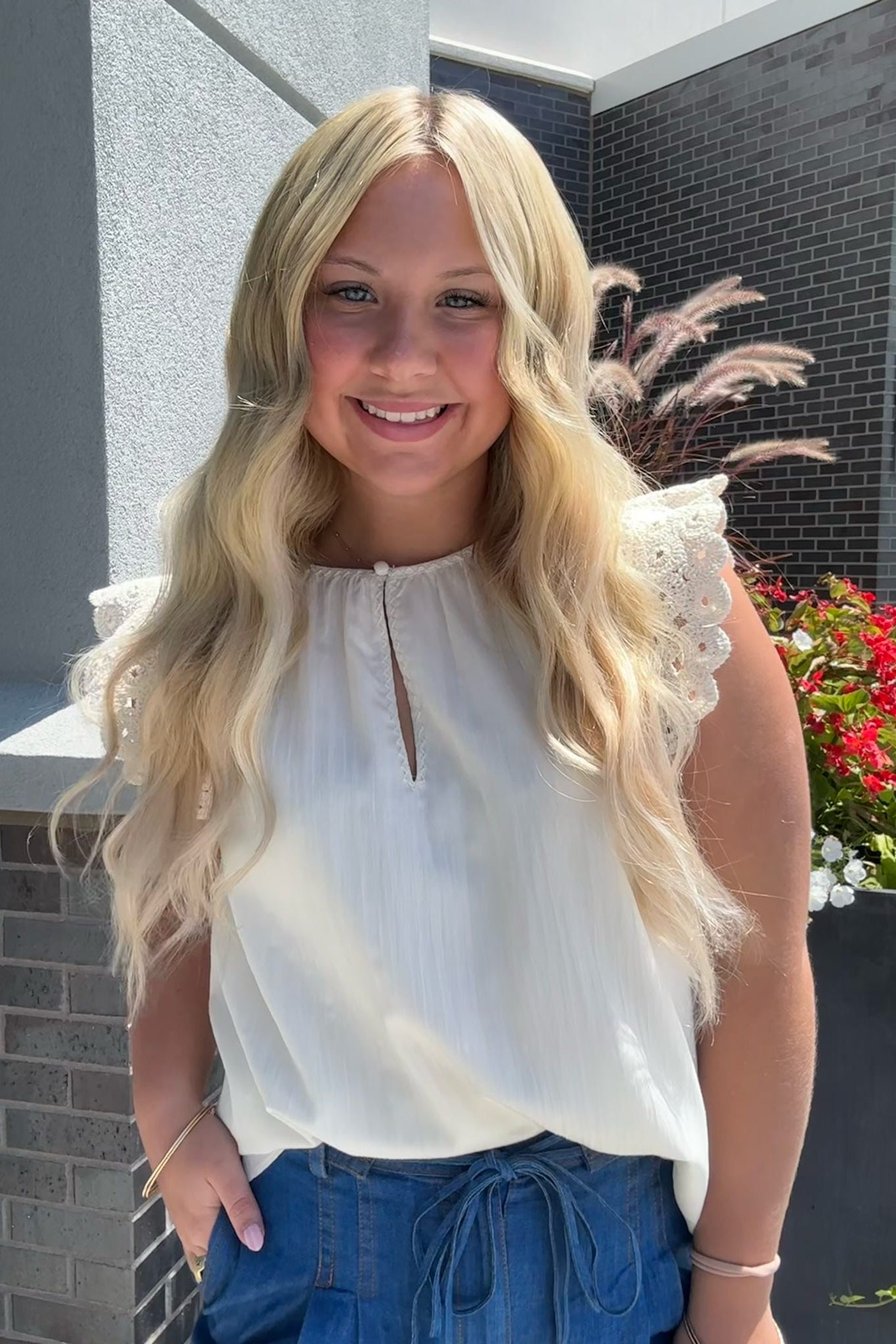 Young woman with long blonde hair wearing a white blouse and blue pants standing outdoors.
