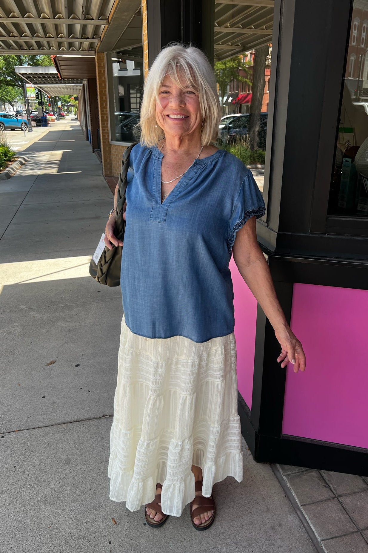 A smiling woman stands on a sidewalk wearing a blue short-sleeve V-neck top and a long cream ruffled skirt, with brown sandals and a shoulder bag. The photo is taken outdoors in front of a storefront with a bright pink panel.
