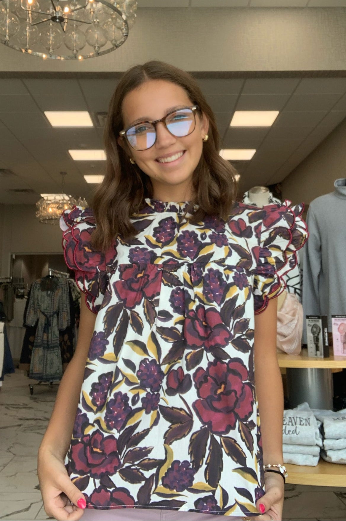 A woman wearing a white short-sleeve blouse with a dark red and purple floral print and puff sleeves, standing inside a clothing store.