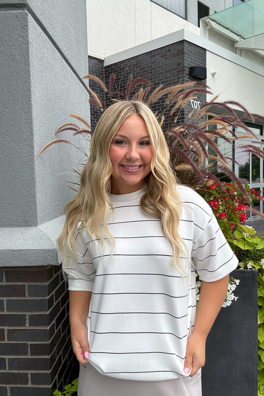 A smiling blonde woman wearing a white short-sleeve t-shirt with thin black horizontal stripes, standing outdoors in front of a modern building with brick walls and potted plants.