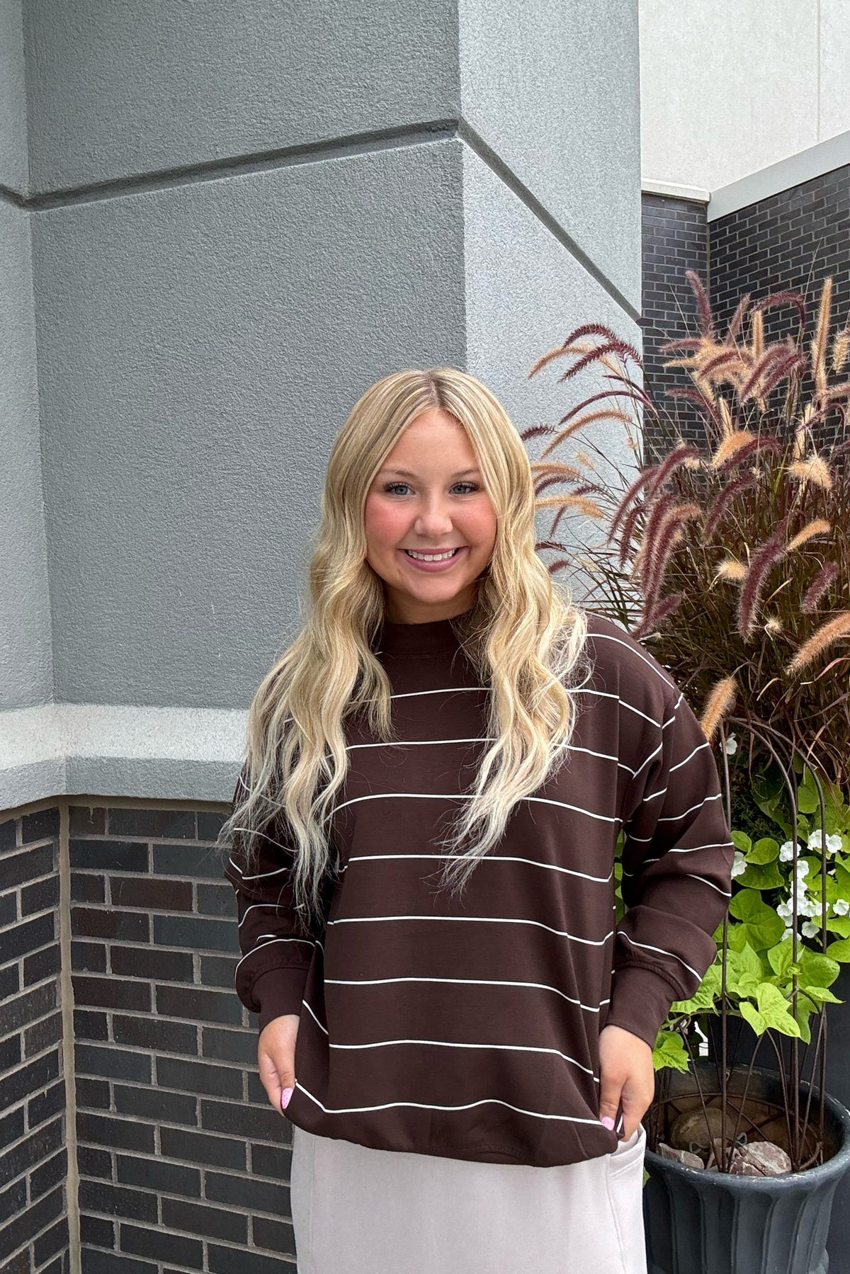 A smiling blonde woman wearing a dark brown mock-neck sweatshirt with thin white horizontal stripes, standing outdoors in front of a gray brick wall and potted plants.