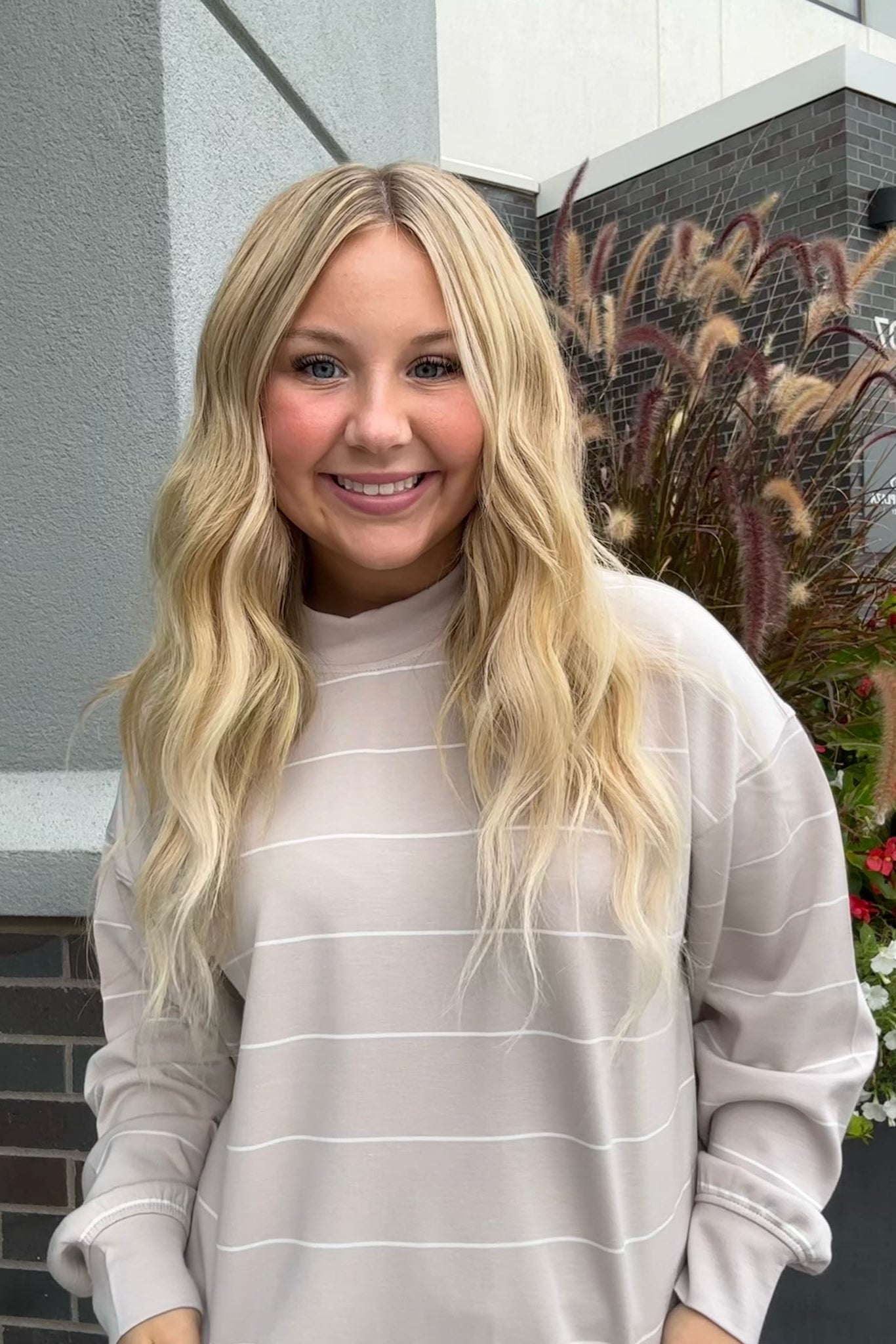 A smiling blonde woman wearing a light beige long-sleeve crewneck sweatshirt with thin white horizontal stripes, standing outdoors in front of a modern building wall with plants.