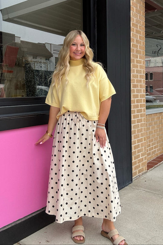 A woman stands on a sidewalk in front of a storefront, wearing a pale yellow short-sleeve mock-neck top and a cream midi skirt with black polka dots. The storefront features a bright pink panel and a tan brick column.