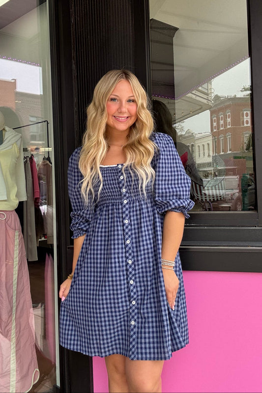 A woman with long blonde hair stands in front of a storefront window wearing a blue and white gingham mini dress with puff sleeves and a button-down front.