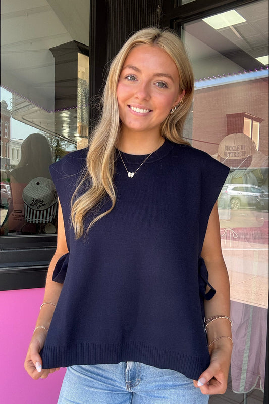 A smiling blonde woman wearing a navy blue sleeveless knit sweater with side tie details and light blue jeans, standing in front of a storefront window.