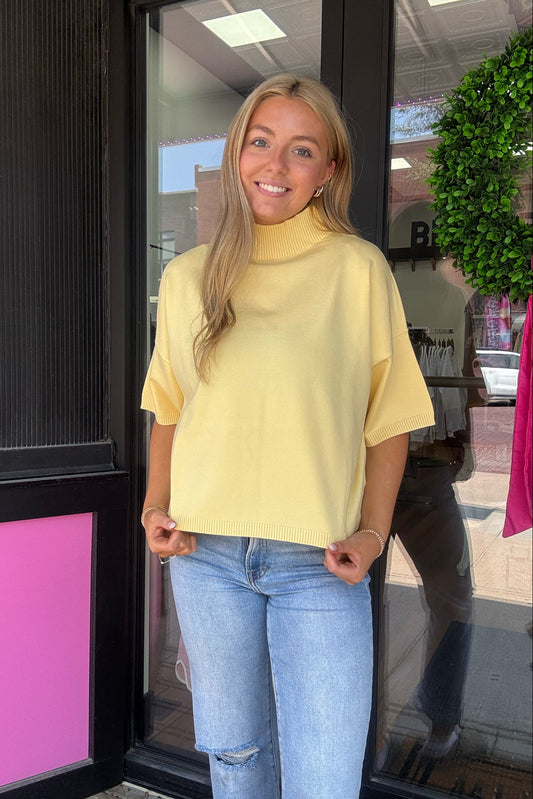 A woman with blonde hair stands in front of a storefront window wearing a pale yellow short-sleeve mock neck knit sweater and light blue jeans.