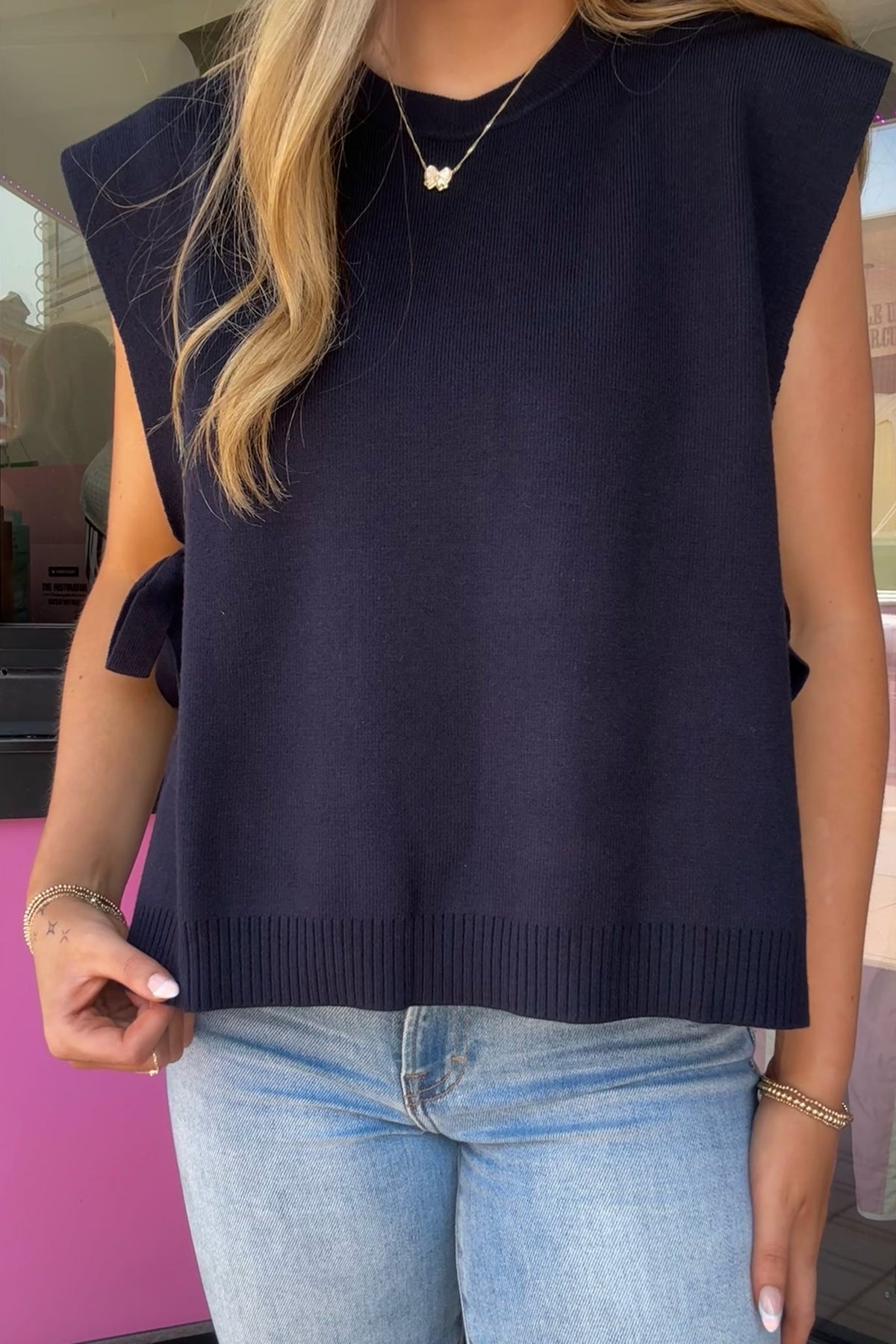A woman wearing a sleeveless navy blue knit sweater vest with side tie details and light blue jeans, shown from the neck down in an outdoor setting.