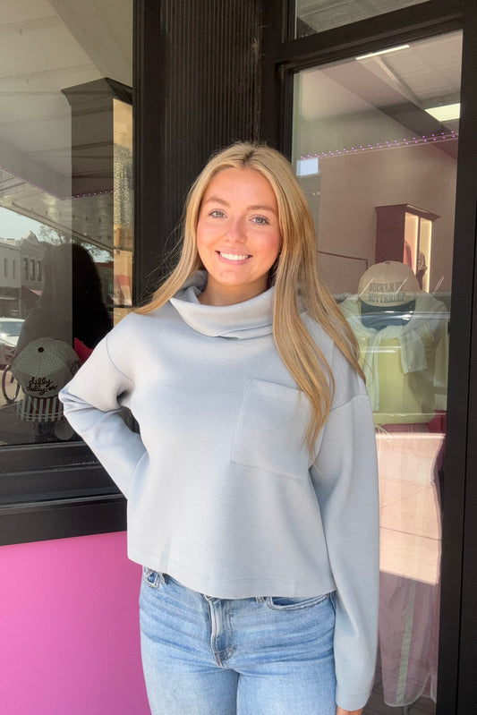 A smiling blonde woman wearing a light blue-gray long-sleeve turtleneck sweater and blue jeans, standing in front of a storefront window.