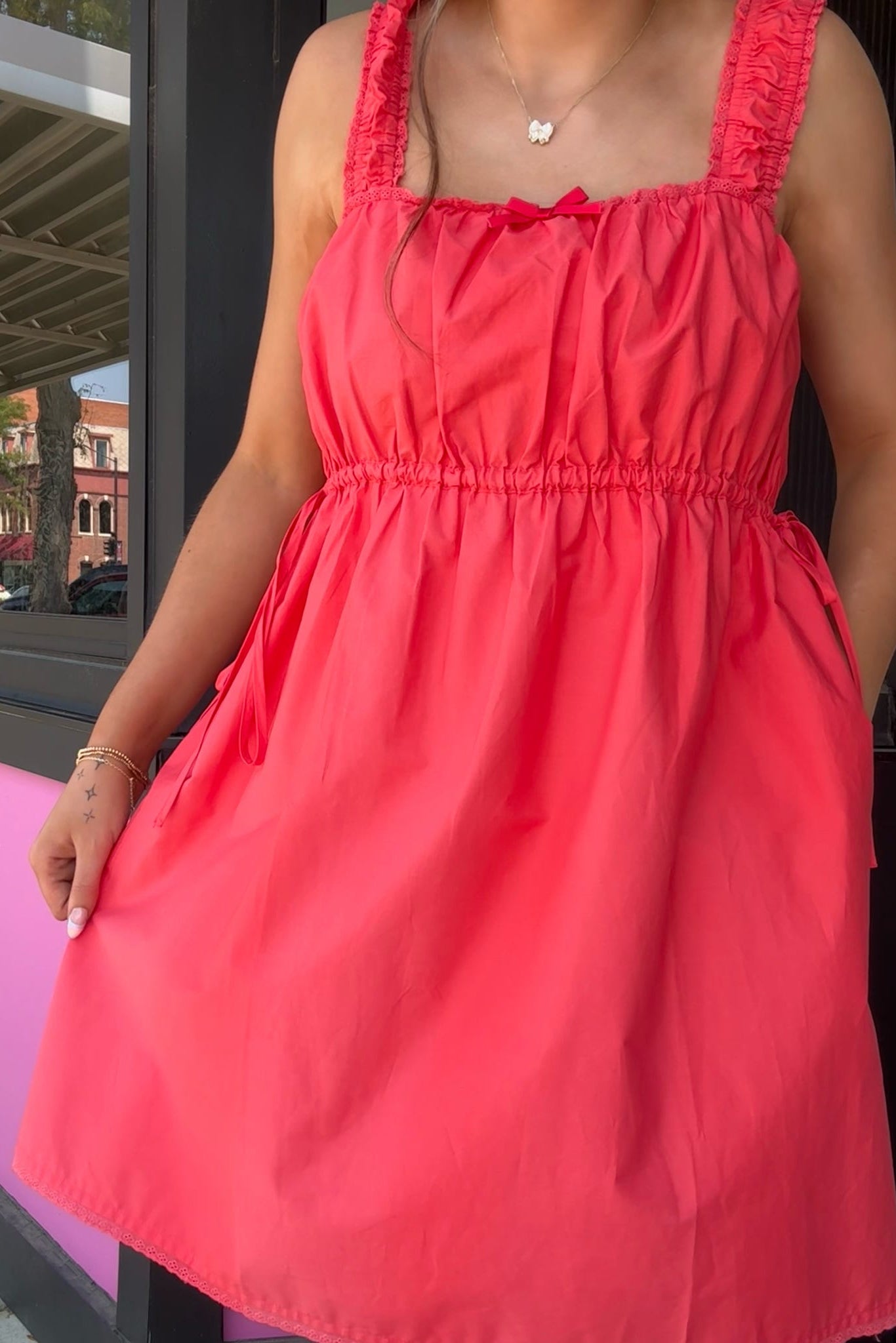 A woman wearing a bright coral pink sleeveless dress with ruffled straps, a square neckline, and a gathered elastic waist, standing in front of a storefront window.