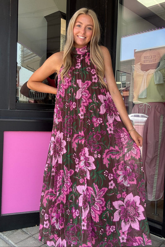 A woman wearing a sleeveless halter-neck maxi dress with a dark brown base and a vibrant pink and purple floral pattern, standing in front of a storefront with pink and black trim.