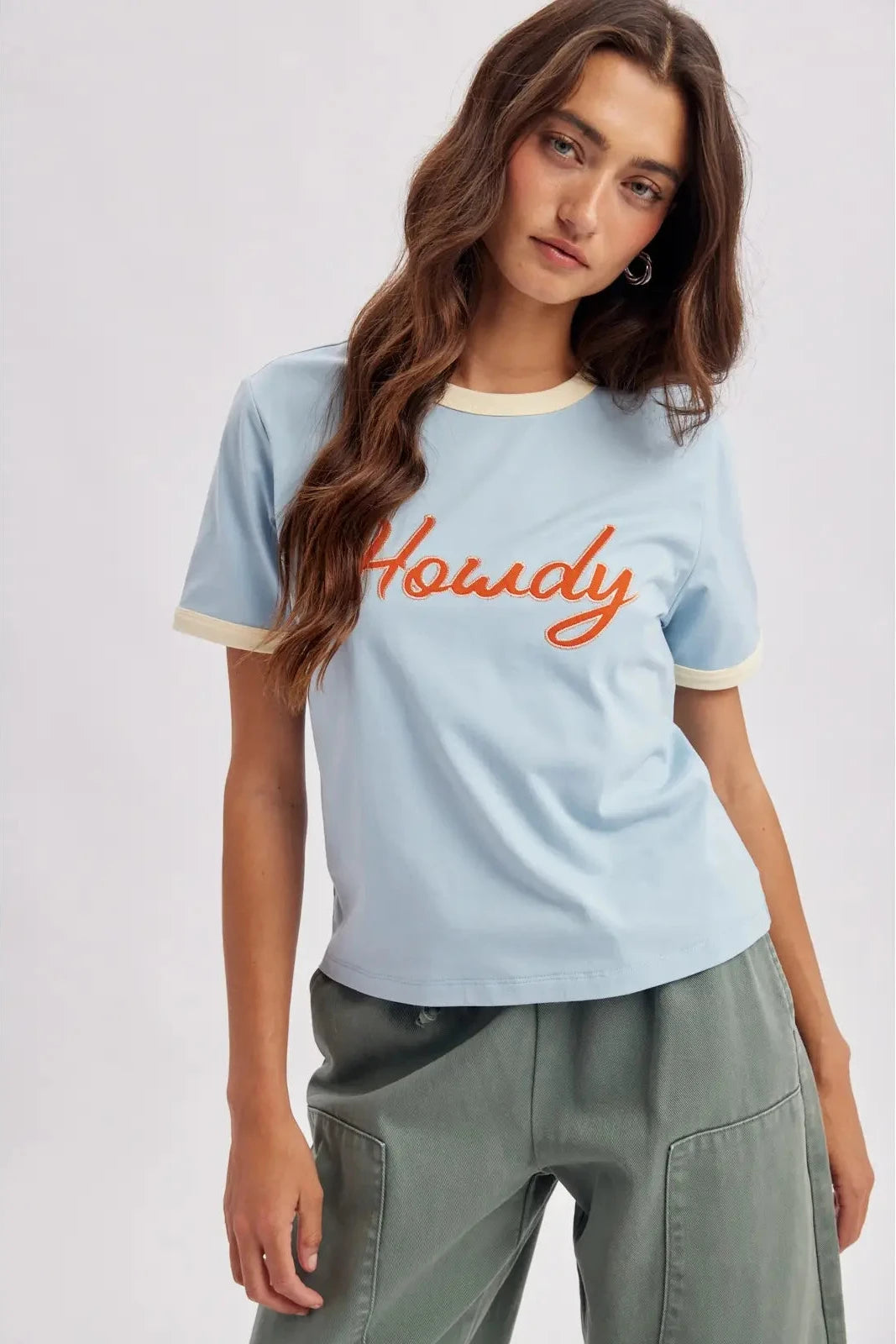 A woman wearing a light blue ringer t-shirt with 'Howdy' embroidered in orange across the chest, paired with sage green cargo pants, against a light gray studio background.