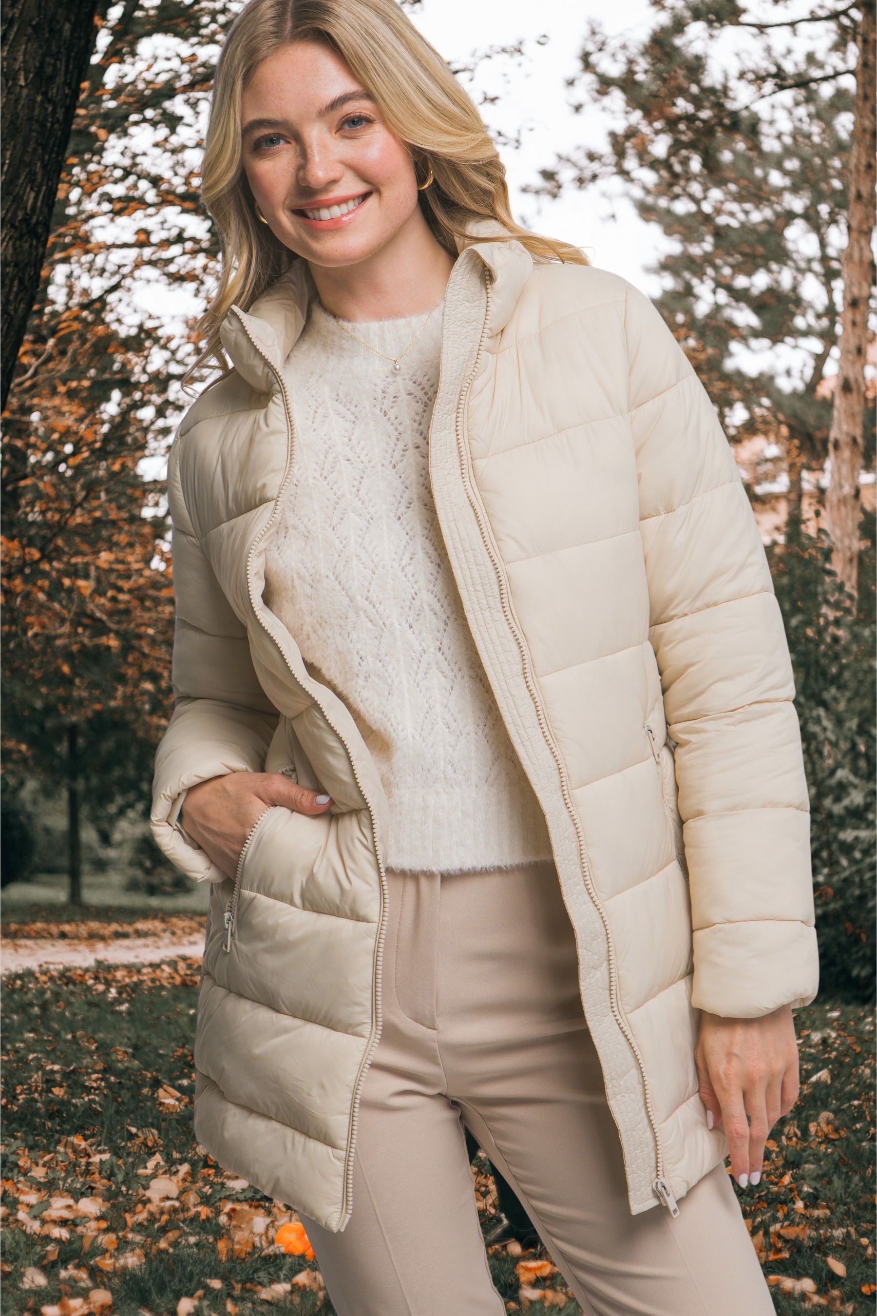 Woman wearing a beige puffer coat in an outdoor setting with trees and fallen leaves.